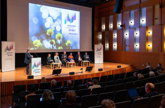 Conference stage where six panelists are sitting and a moderator is standing. In the background there is text Rovaniemi Arctic Spirit.